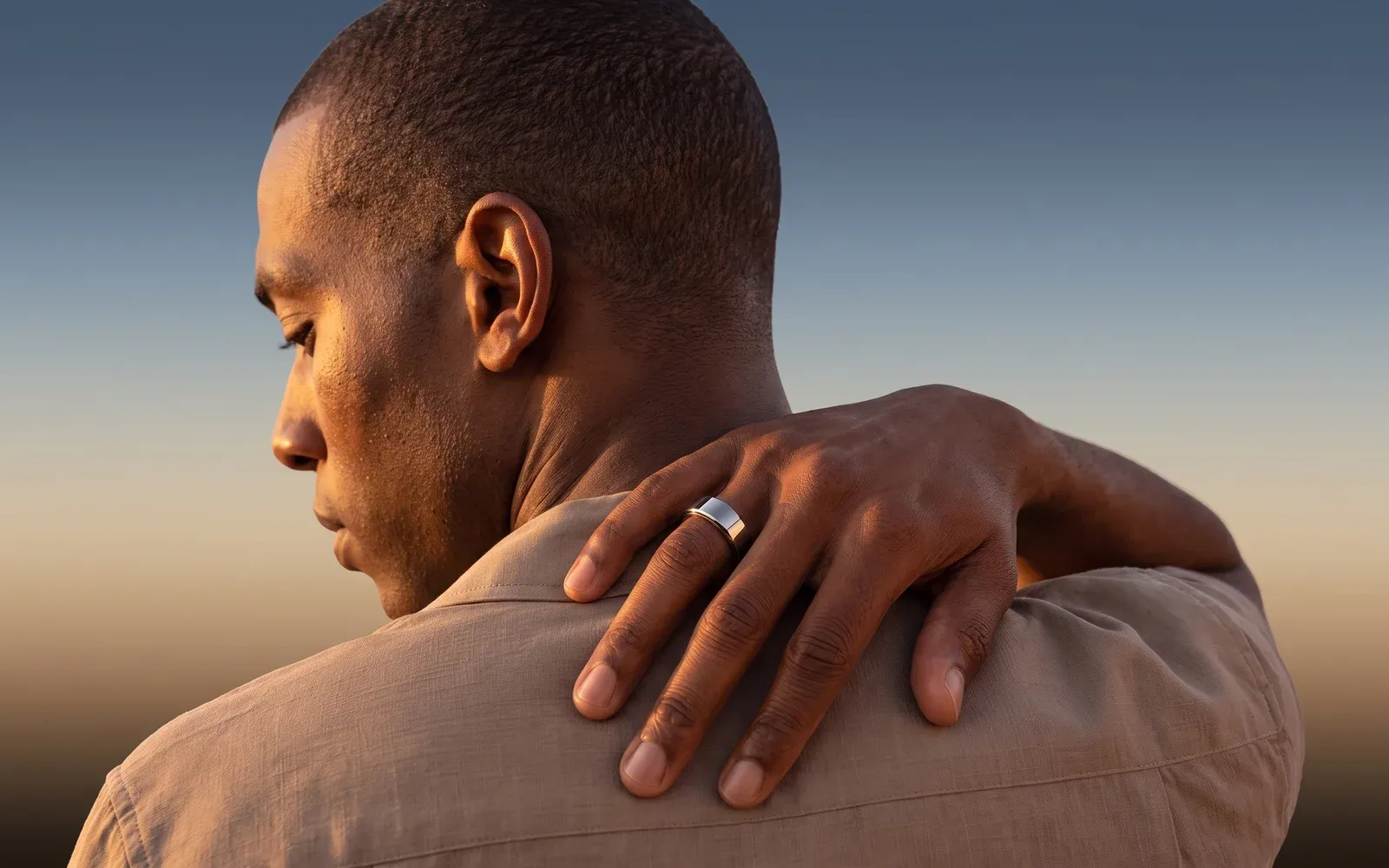A man in a linen shirt looking down, a hand wearing the silver Ain ring on his shoulder