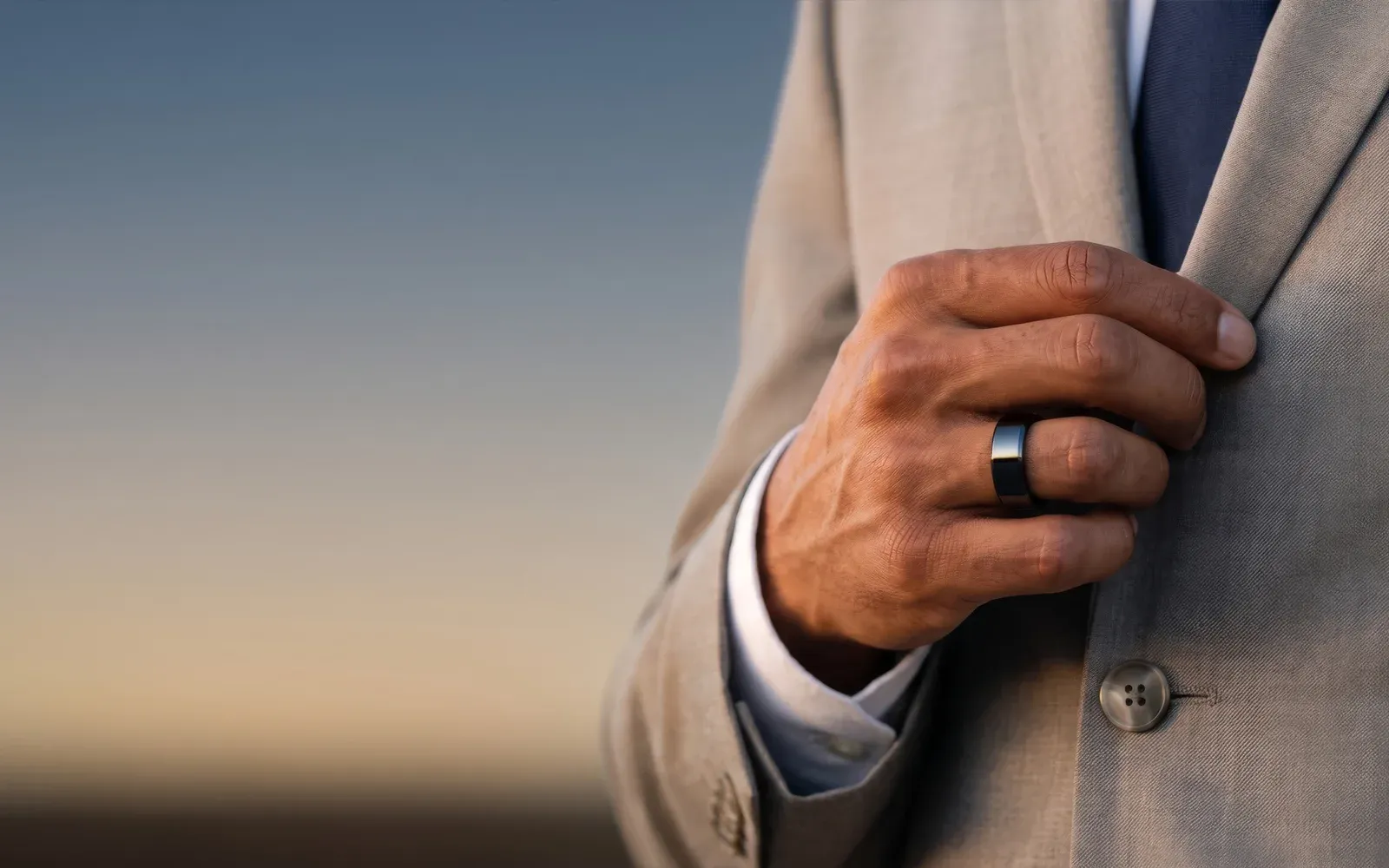 A man in a sand-coloured suit holding his lapel, the black Hasa ring on his finger