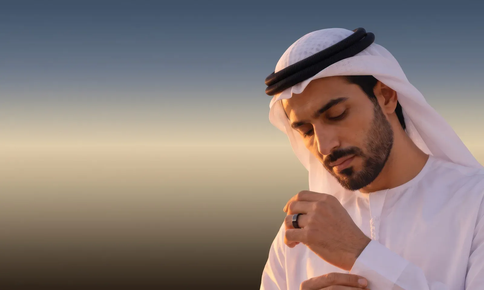 A man in a white kandura and ghutra looking down at his cuff, the black Hasa ring on his finger