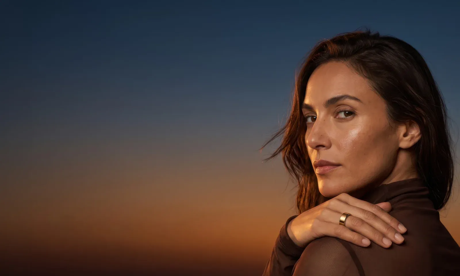 A woman looking back over her shoulder at dusk, her hand on her shoulder wearing the gold Liwa ring