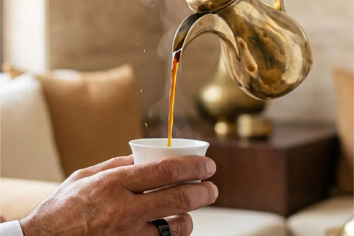 Arabic coffee poured from a brass dallah into a small cup, held by a hand wearing a dark ring
