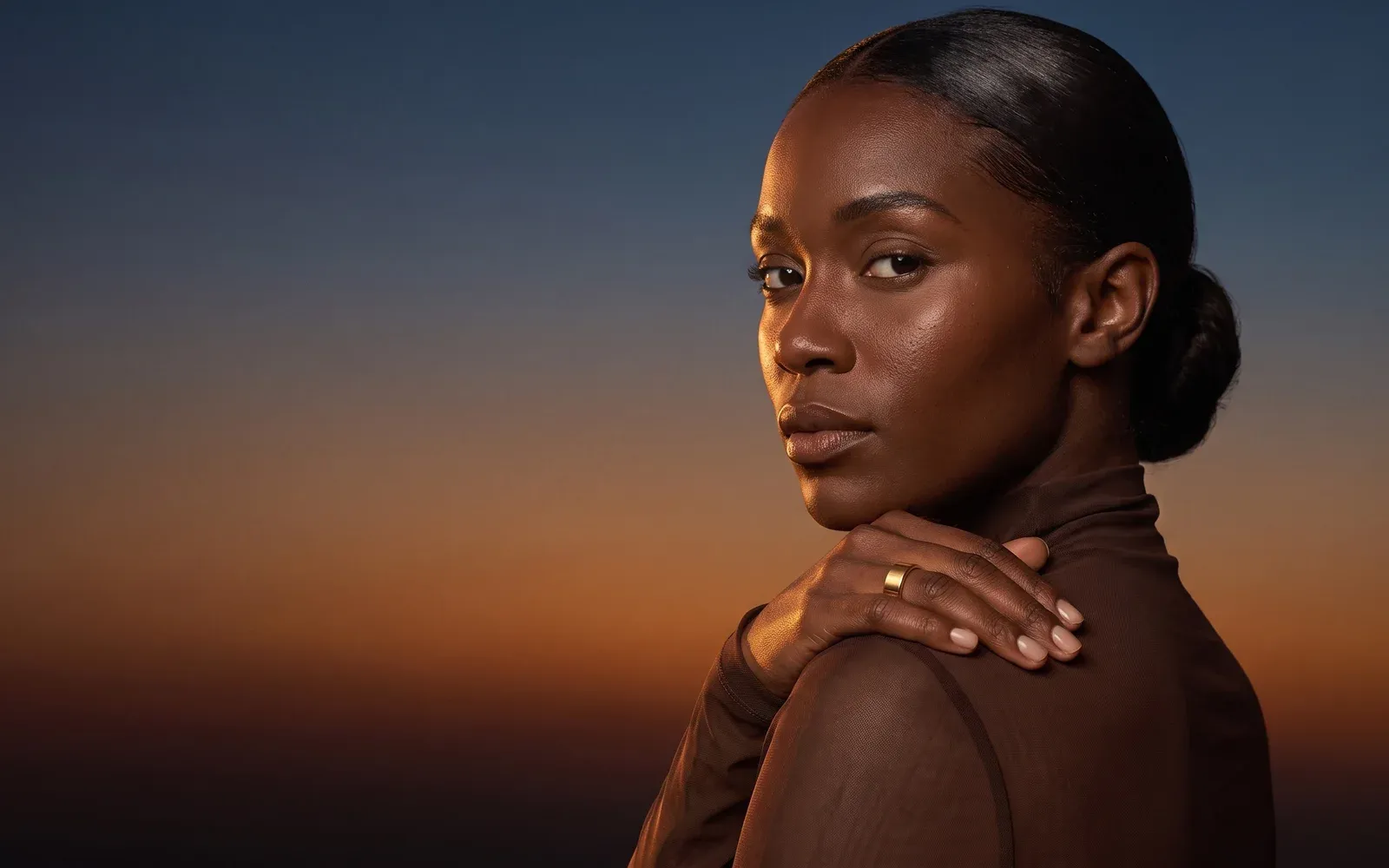 A woman with her hair tied back, her hand on her shoulder wearing the gold Liwa ring, the sky at dusk behind